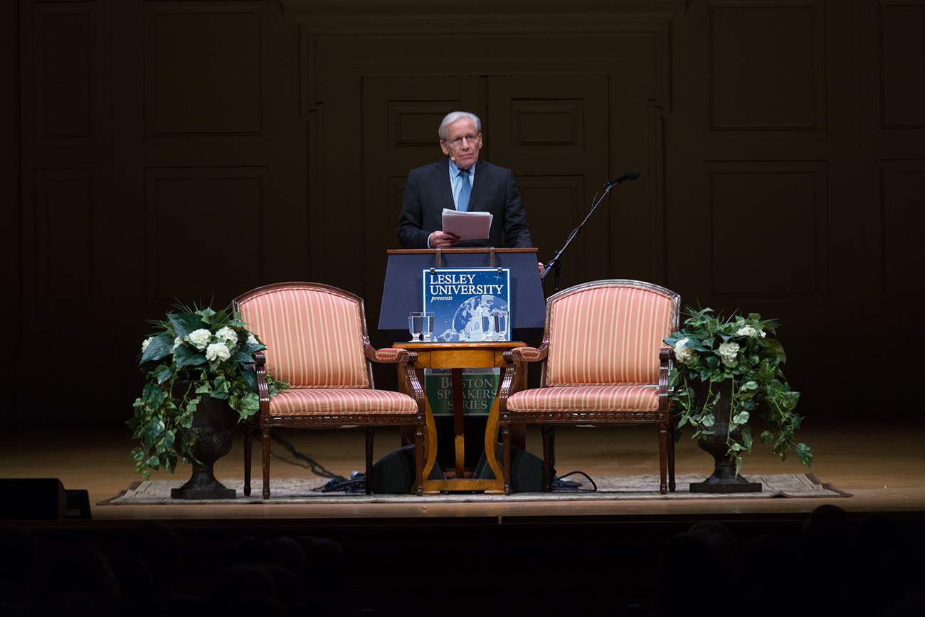 A man standing behind a podium giving a speech between two chairs on a dark stage.