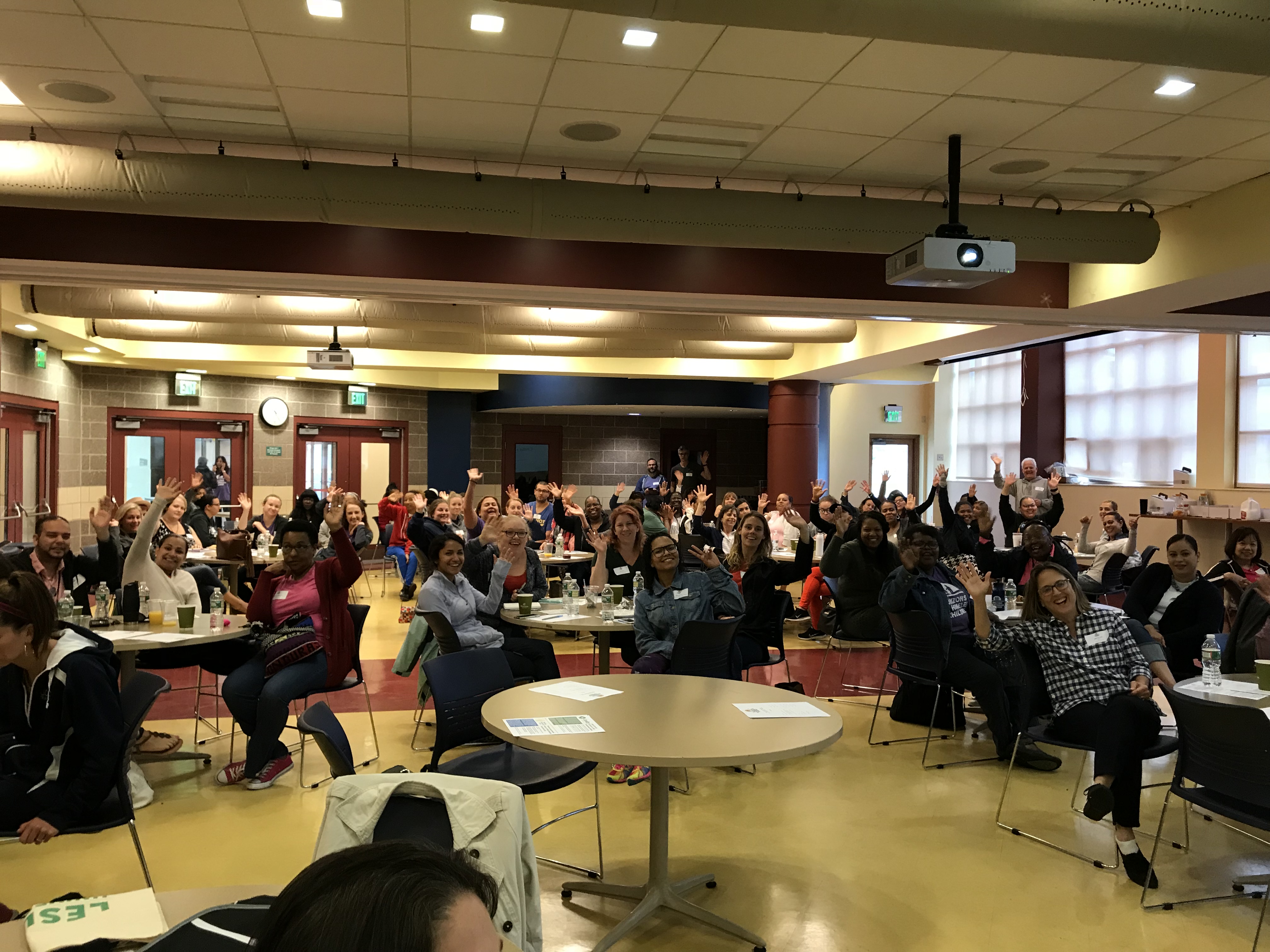 A group of people in a school cafeteria at round tables, waving. 