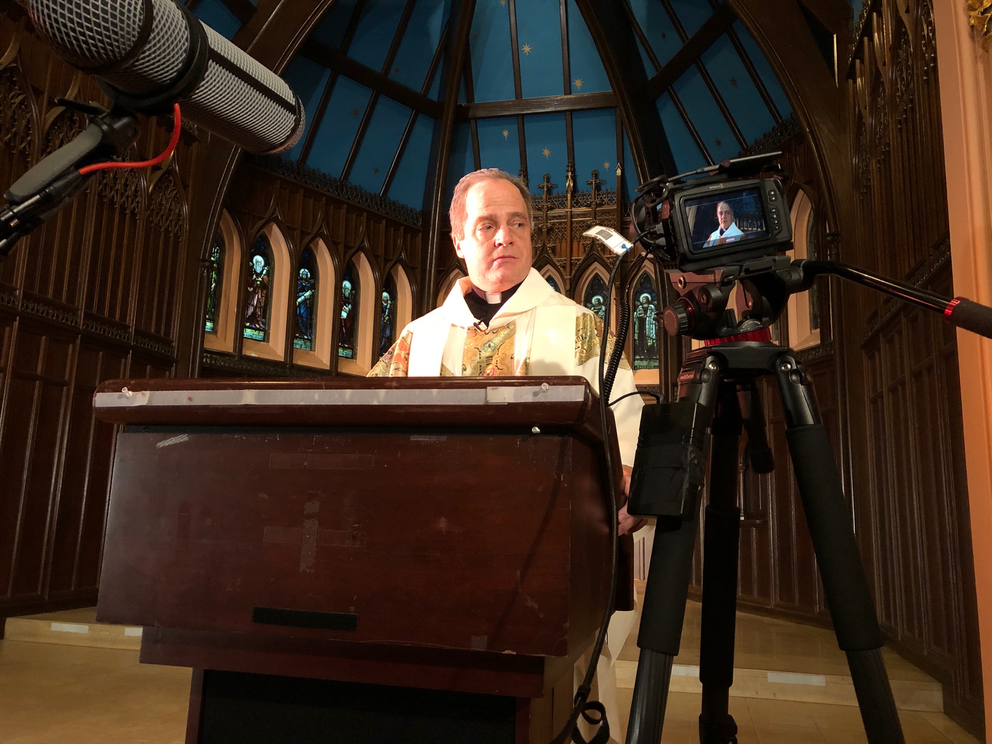 Scene from "Stay" - a man dressed as a priest at an altar with video cameras visible