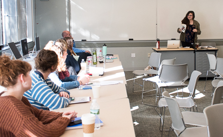 Jess Rizkallah stands at the head of the room speaking to students who are seated to the left of the image.