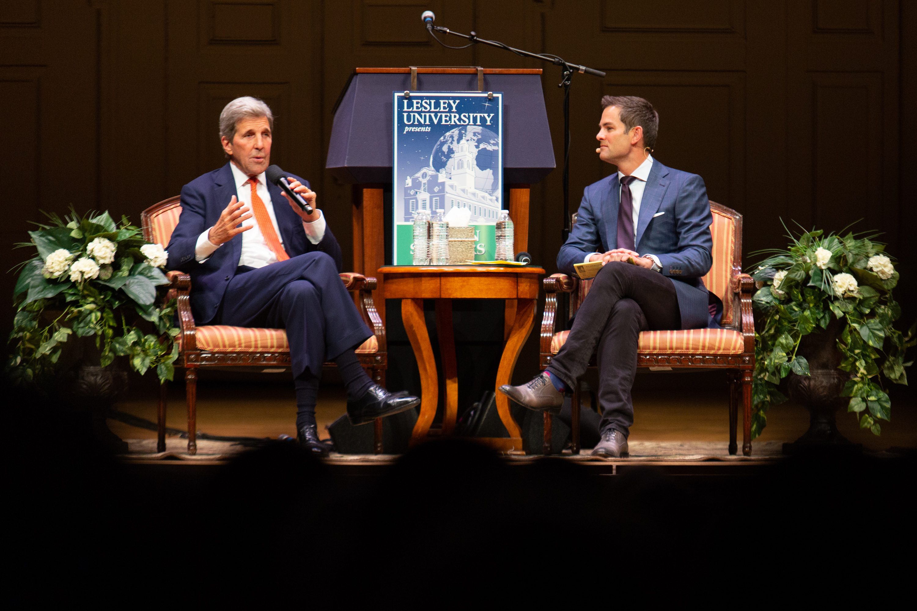 John Kerry and Jared Bowen are seated on the Symphony Hall stage with flowers on either side