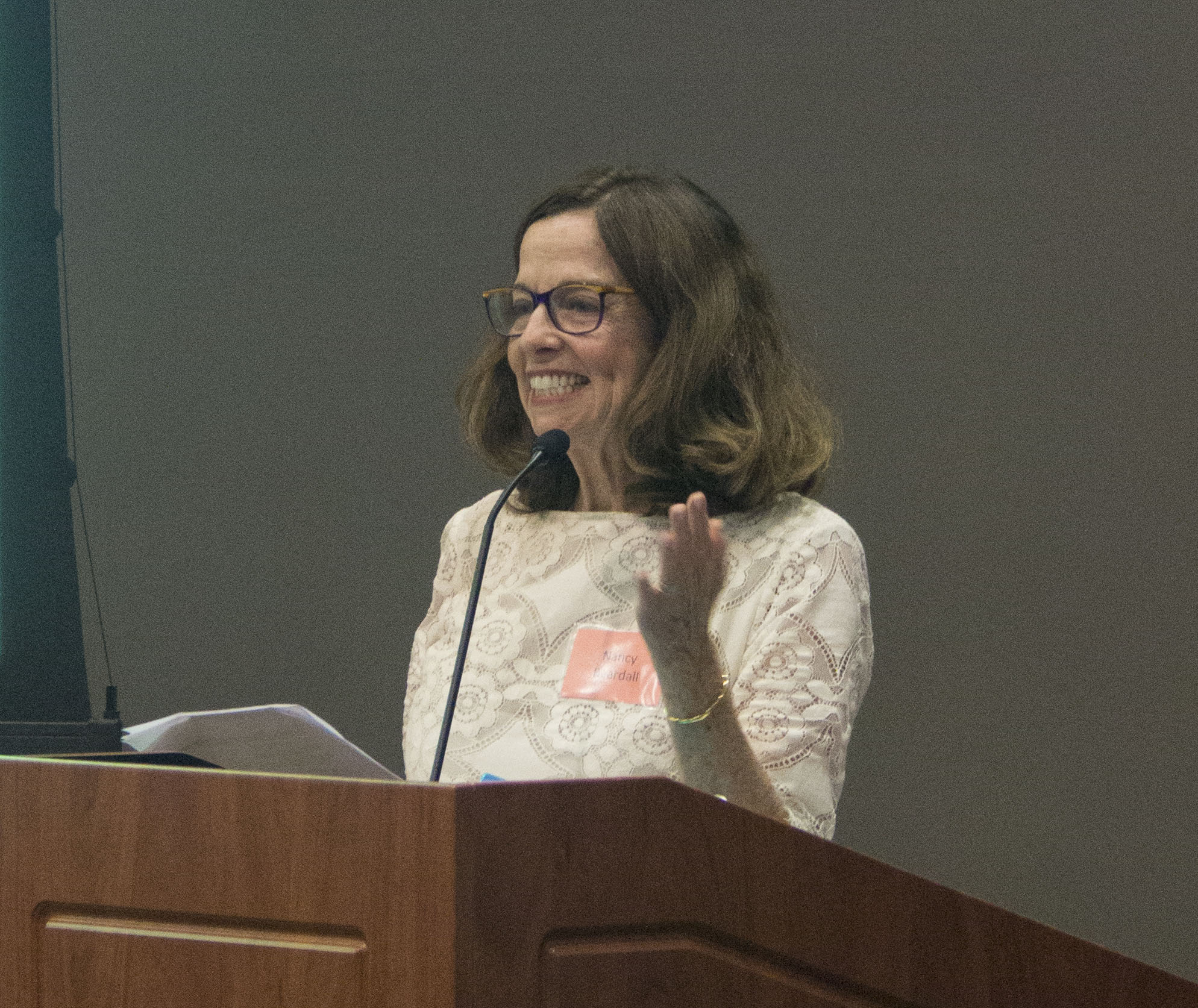 Woman with brown hair and glasses behind a podium, speaking into a microphone