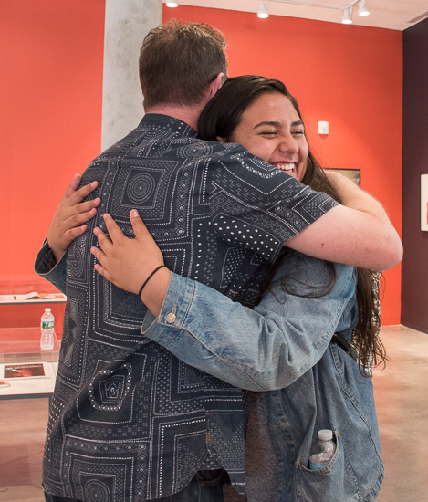 Standing in the art gallery, Vaughan Oliver hugs a female student who has a smile on her face.