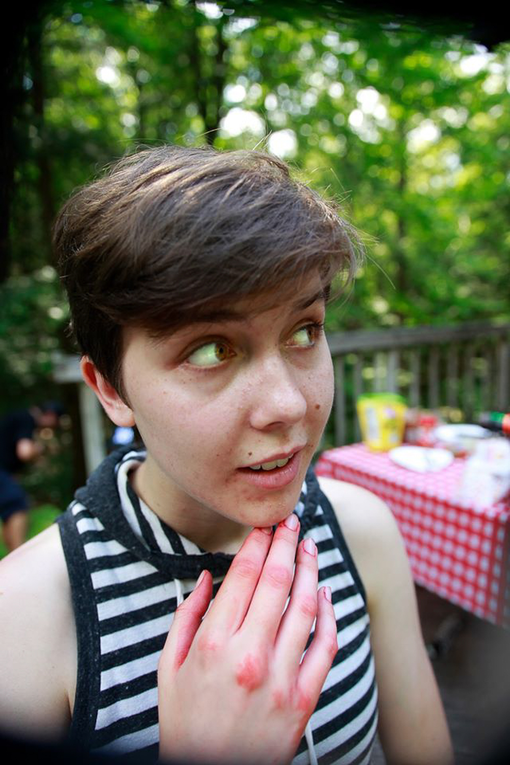 Person with short sleeved t-shirt on and hand just under their chin, hand has fake blood residue