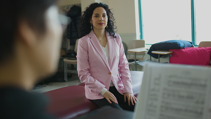 Woman in a pink blazer sitting on a bench in a classroom behind a woman playing the piano with sheet music. 