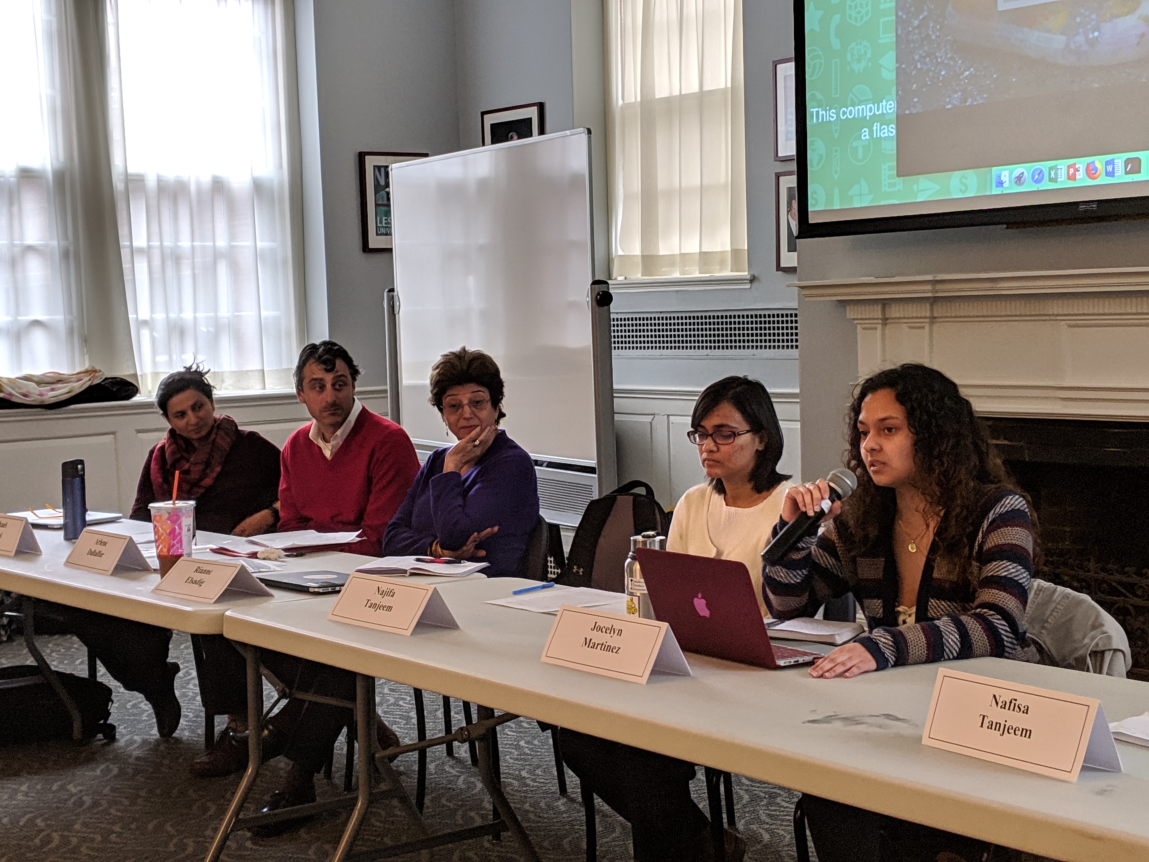 A panel of 5 speakers behind a table in Alumni Hall in front of a screen.