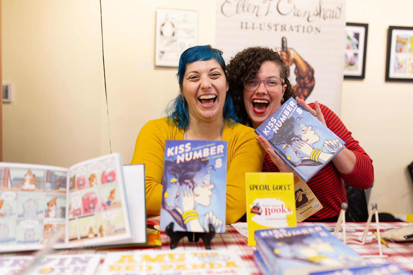 Ellen and Colleen sitting behind a table with their books on it, sitting next to each other laughing and holding their books up to their face