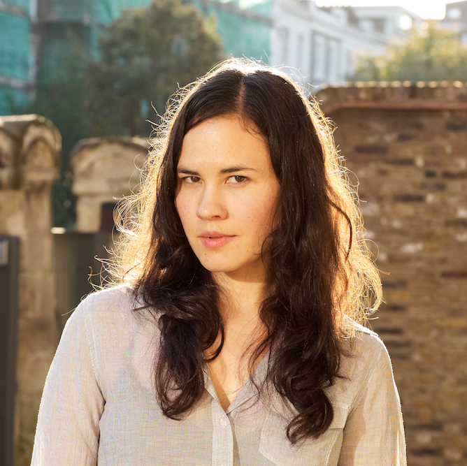 A photograph of Laurel Nakadate looking strait at the camera with a stone wall behind her. The sun illuminates her hair from behind. 