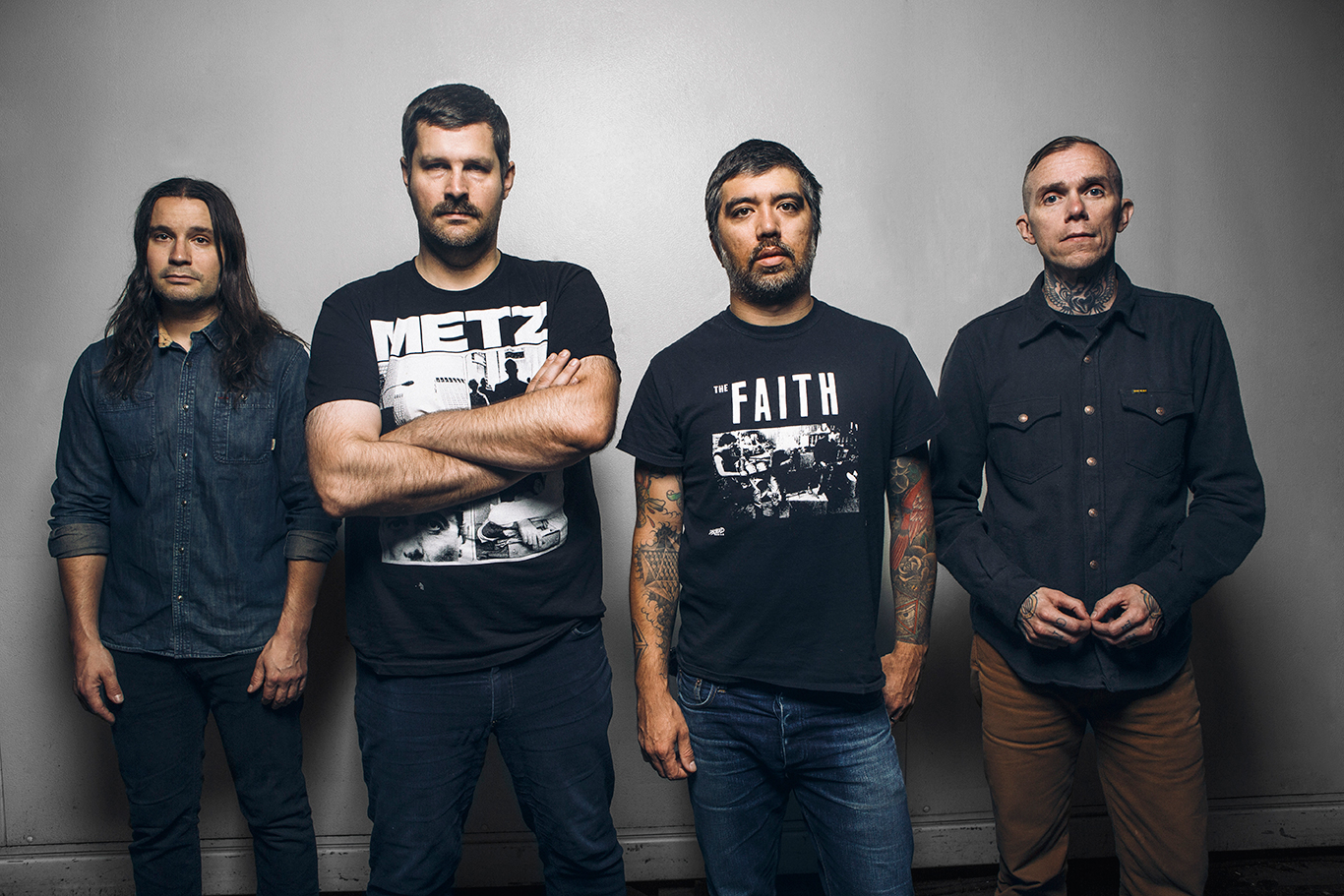 four men stand against a gray wall in black t-shirts 