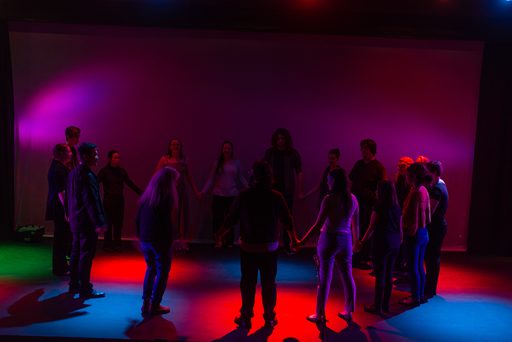 A group of actors in a circle on stage in Marran theater with dark, colorful lighting