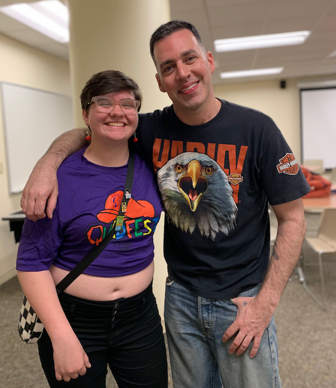 Student in purple t-shirt posing with tall man in black t-shirt in University Hall classroom