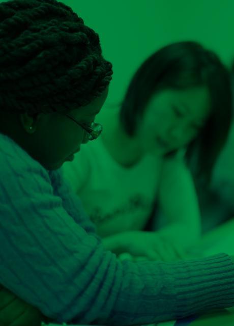 All green image of graduate students in class. Two women are reading text. Three other students in the background look forward.