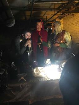 Three guys in a basement stand around a table wearing medevil knight costumes holding swords.