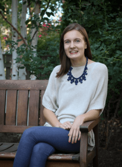 Helen smiles while sitting on a wooden bench outside. Helen wears a white sweater, blue jeans, and a blue necklace.