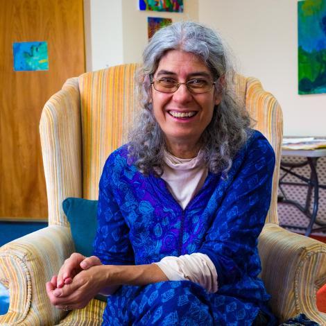 Photo of Jennie Kristel sitting in chair with hands folded in her lap, smiling at camera. She is wearing a royal blue shirt sitting on a neutral colored chair. 