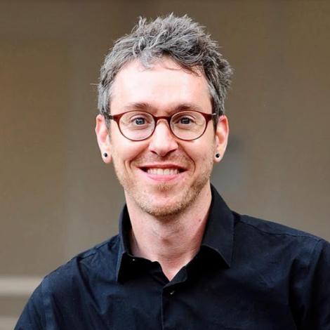 Headshot Darrell Penta smiling, wearing a navy blue collared shirt, black earrings, and rounded glasses.