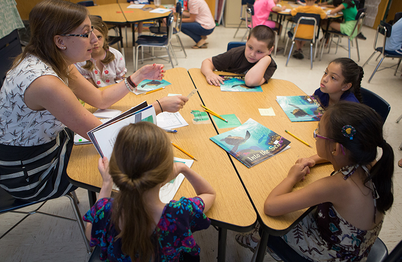 A group of students sitting at a table reading books with a teacher